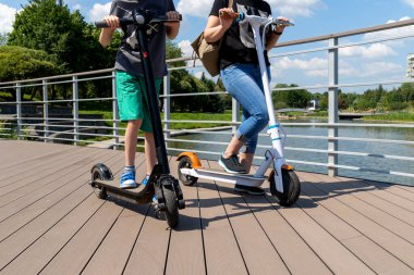 Genç bir çocuk ve bir kadın parkta iki elektrikli scooter kullanıyor. Güneşli bir yaz günü. Modern şehir taşımacılığı. Nehrin kenarındaki şehir parkı.