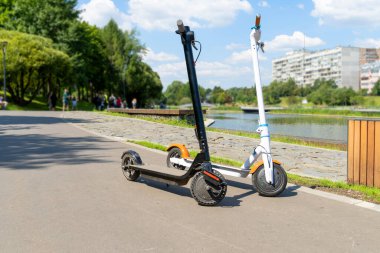 İki elektrikli scooter, siyah ve beyaz, caddedeki tramvayda duruyor. Güneşli bir yaz günü. Modern şehir taşımacılığı. Nehrin kenarındaki şehir parkı.