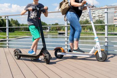 Genç bir çocuk ve bir kadın parkta iki elektrikli scooter kullanıyor. Güneşli bir yaz günü. Modern şehir taşımacılığı. Nehrin kenarındaki şehir parkı.