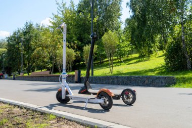 İki elektrikli scooter, siyah ve beyaz, caddedeki tramvayda duruyor. Güneşli bir yaz günü. Modern şehir taşımacılığı. Ağaçlı şehir parkı.