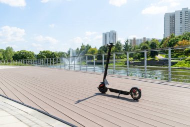 Elektrikli siyah bir scooter caddedeki tramvayda duruyor. Şehir parkında korkulukların olduğu ahşap döşemeler var. Güneşli bir yaz günü. Modern şehir taşımacılığı.