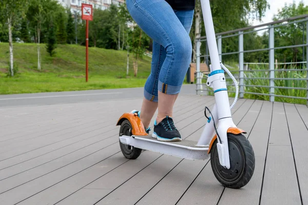 Kot pantolonlu bir kız sokakta beyaz elektrikli scooter kullanıyor. Şehir parkı, yaz güneşli bir gün, ahşap gezinti güvertesi. Modern şehir taşımacılığı. Scooter 'a binen kadın.