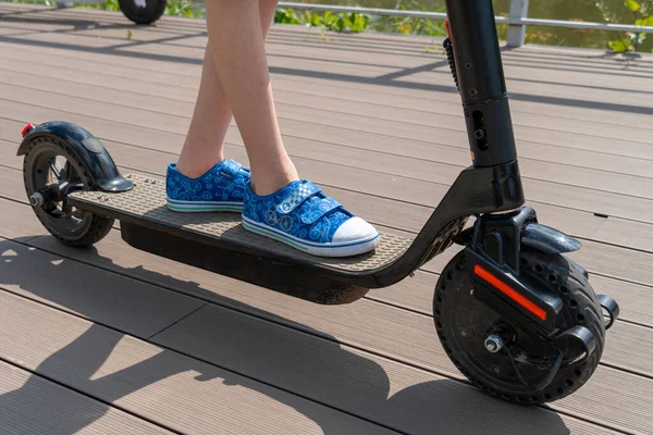 Genç bir çocuk sokakta elektrikli scooter kullanıyor. Şehir parkı, yaz güneşli bir gün, nehir kenarındaki ahşap gezinti alanı. Modern şehir taşımacılığı. Yeşil şortlu bir çocuk scooter sürüyor..