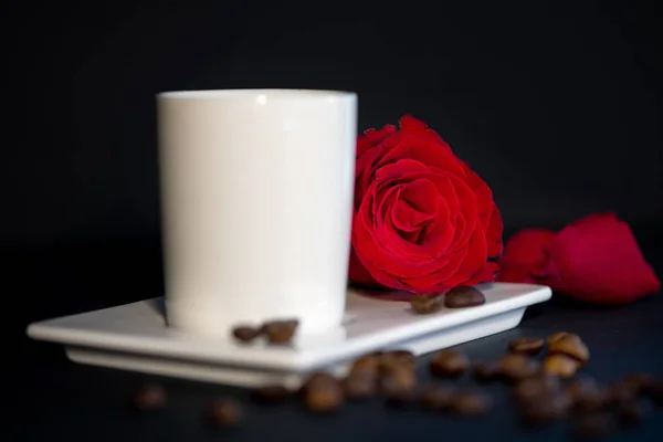 white cup of coffee, red rose, coffee beans and rose petals on a dark background