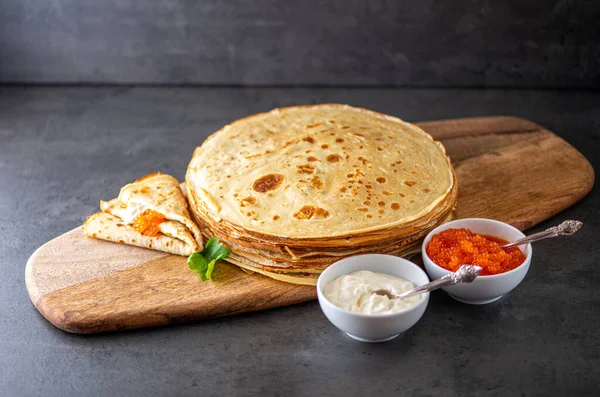 Bir yığın ev yapımı krep ahşap bir tahta üzerinde siyah bir arka plan ve bir rulo krep, ve onun üzerinde kırmızı havyar ve ekşi krema, ve yanında ekşi krema ile beyaz kaseler