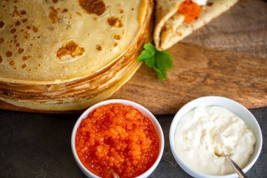 Bir yığın ev yapımı krep ahşap bir tahta üzerinde siyah bir arka plan ve bir rulo krep, ve onun üzerinde kırmızı havyar ve ekşi krema, ve yanında ekşi krema ile beyaz kaseler