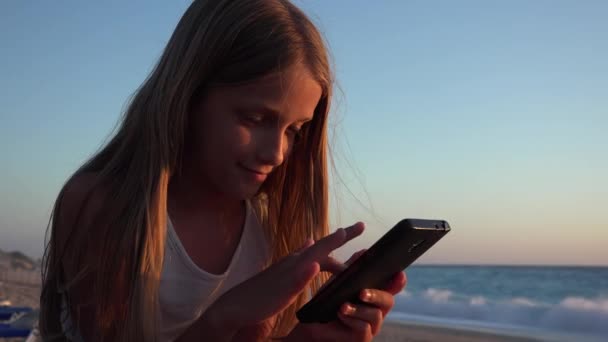 Ребенок играет в смартфон, ребенок на пляже на закате, девушка с планшета на берегу моря — стоковое видео