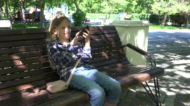 Kid Playing Smartphone Przeglądanie Internetu na placu zabaw w parku, Dziewczyna siedzi na ławce, Dziecko na świeżym powietrzu w parku — Wideo stockowe