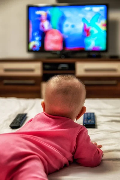 Lying girl infant with remote controllers and watching cartoons on TV screen