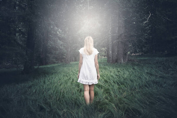 Back view of a blonde woman in white summer dress in mystic forest with grass floor. Concept spiritual.