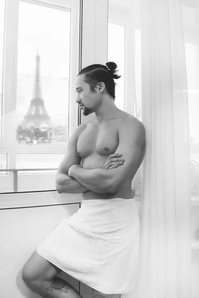 Bonito, jovem musculoso em toalha branca enrolada em torno de sua cintura olhando pela janela pela manhã — Fotografia de Stock
