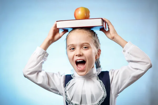 Feliz colegiala con libro y manzana roja en la cabeza, vistiendo blusa blanca vintage y gafas de montura negra, de pie sobre fondo azul. Regreso al concepto escolar —  Fotos de Stock