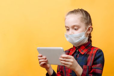 Sarı arka planda tıbbi maskeli genç bir kız elinde bir tablet bilgisayar tutuyor..