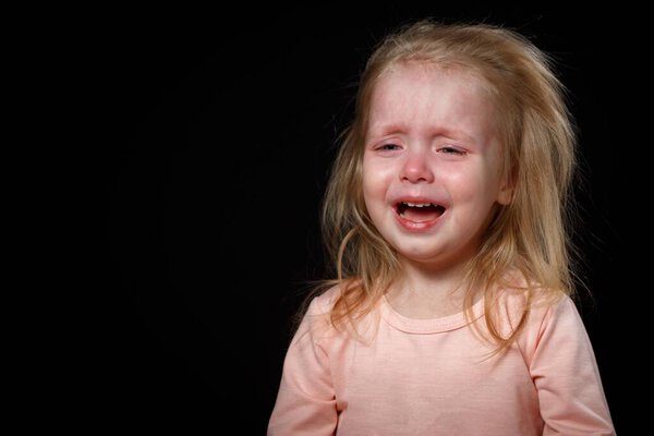 Маленькая девочка громко плачет, она очень расстроена, она истерична. Она не может успокоиться. Фото в студии на темном фоне
.