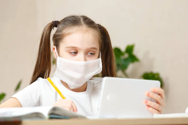 Distance learning online education. Schoolgirl girl in medical mask with white digital tablet at home studying lessons. Online concept of school education and social distance during quarantine, self-isolation