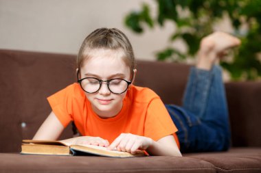 Gözlüklü, sıradan kıyafetleri olan, kanepede uzanmış kitap okuyan beyaz bir kız.