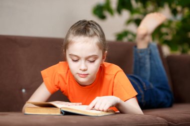 Koronavirüs yüzünden karantina sırasında evdeki kanepede uzanmış kitap okuyan sevimli beyaz bir kız. Eğitim ve eğlence kavramı.