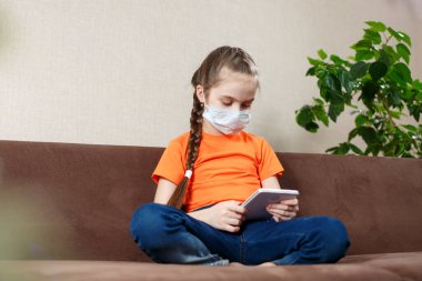 Maskeli küçük kız Lotus pozisyonunda kanepede oturuyor ve tablet bilgisayar kullanıyor. Evde yalnızlık