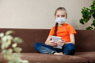 Maskeli küçük kız Lotus pozisyonunda kanepede oturuyor ve tablet bilgisayar kullanıyor. Evde izolasyon. Kameraya bakıyorum..