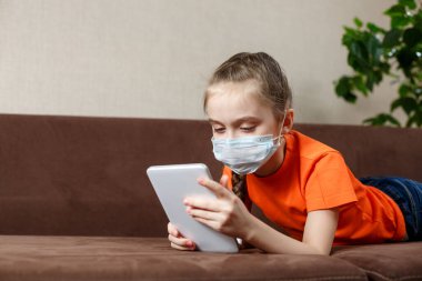 Maskeli küçük kız kanepede uzanıyor ve tablet bilgisayar kullanıyor. Evde yalnızlık