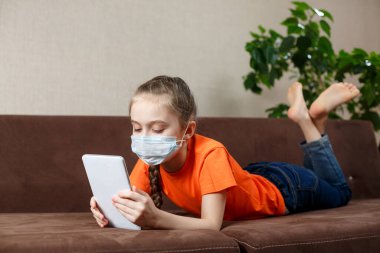 Maskeli küçük kız kanepede uzanıyor ve tablet bilgisayar kullanıyor. Evde yalnızlık