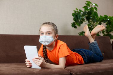 Maskeli küçük kız kanepede uzanıyor ve tablet bilgisayar kullanıyor. Evde izolasyon. Kameraya bakıyorum.