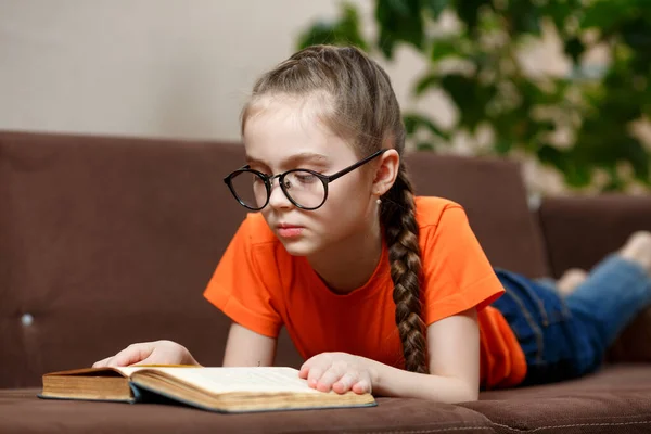 Cute Kaukaska Dziewczynka Okularach Długim Warkoczem Czytając Książkę Leżąc Kanapie — Zdjęcie stockowe