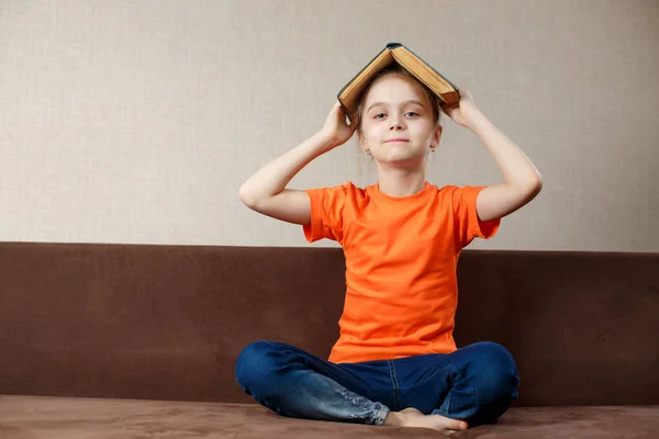 Gelukkig Schoolmeisje Met Boek Haar Hoofd Oranje Shirt Jeans Zittend — Stockfoto