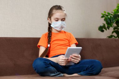 Maskeli küçük kız Lotus pozisyonunda kanepede oturuyor ve tablet bilgisayar kullanıyor. Evde yalnızlık