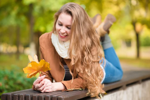 Beauty woman holding autumn leaves and relaxing a bench — Stock Photo, Image