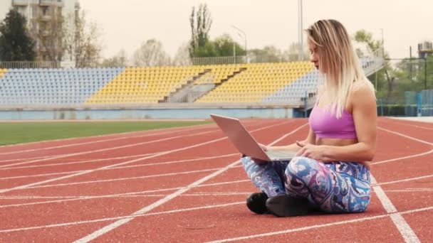 Mädchen beim Online-Workout mit Laptop im leeren Stadion. Charmante Blondine. Während der Quarantäne. — Stockvideo