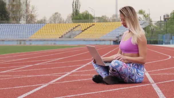 Chica haciendo ejercicio en línea con el ordenador portátil en el estadio vacío. Una rubia encantadora. Durante la cuarentena . — Vídeo de stock