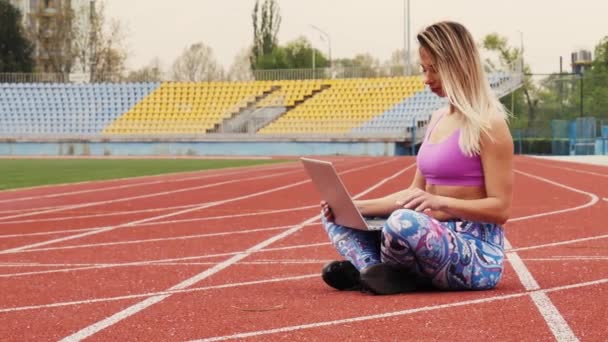 Девушка занимается онлайн тренировкой с ноутбуком на пустом стадионе. Прекрасная блондинка. Во время карантина . — стоковое видео