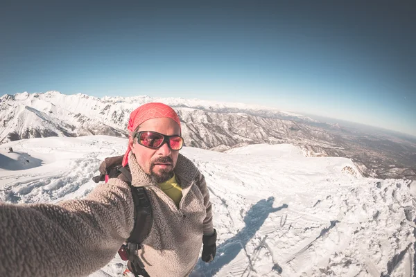 Adult alpin skier with beard, sunglasses and hat, taking selfie on snowy slope in the beautiful italian Alps with clear blue sky. Toned image, vintage style, ultrawide angle fisheye lens. — Stock Photo, Image