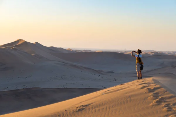 Tourist holding smart phone and taking photo at scenic sand dunes illuminated by sunset light in the Namib desert, Namib Naukluft National Park, Namibia. Adventure and exploration in Africa. — Stock Photo, Image