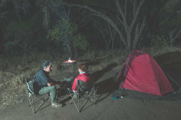 Pareja adulta relajándose en camping por la noche. Aventura en el Parque Nacional, Sudáfrica. Quemando fuego y tienda de campaña en el fondo. Imagen tonificada . —  Fotos de Stock