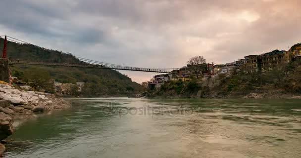 Caducidade do tempo crepúsculo em Rishikesh, cidade santa e destino de viagem na Índia. Céu colorido e nuvens em movimento sobre o rio Ganges. Pessoas que atravessam a ponte suspensa . — Vídeo de Stock
