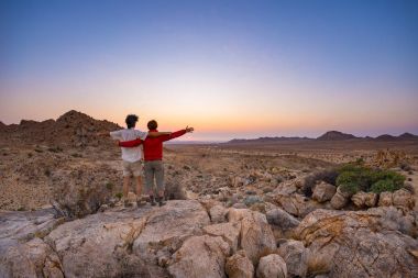 Sarılma çift uzanmış kollarıyla Namib Çölü, Namibya, Afrika görkemli ziyaretçi cazibe çarpıcı görünümü izliyor. Turuncu kırmızı mor açık gökyüzü alacakaranlıkta ufuktan. Peop seyahat