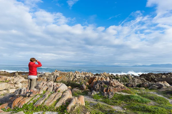 Turista olhando com binocular na linha costeira rochosa em De Kelders, África do Sul, famosa pela observação de baleias. Estação de inverno, céu nublado e dramático . — Fotografia de Stock