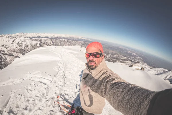 Adult alpin skier with beard, sunglasses and hat, taking selfie on snowy slope in the beautiful italian Alps with clear blue sky. Toned image, vintage style, ultrawide angle fisheye lens. — Stock Photo, Image
