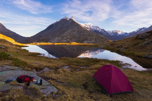 Camping with tent near high altitude lake on the Alps. Reflection of snowcapped mountain range and scenic colorful sky at sunset. Adventure and exploration. — Stock Photo, Image