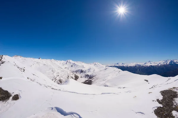 Sonnenstern, der an einem strahlend sonnigen Wintertag über schneebedeckten Bergketten und hohen Berggipfeln im italienischen Alpenbogen leuchtet. offen schneebedeckter Hang im Vordergrund. — Stockfoto