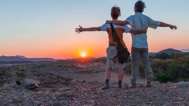 Sarılma çift uzanmış kollarıyla Namib Çölü, Namibya, Afrika görkemli ziyaretçi cazibe çarpıcı görünümü izliyor. Turuncu kırmızı mor açık gökyüzü alacakaranlıkta ufuktan. Peop seyahat