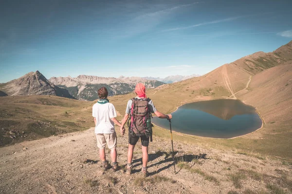 Un par de excursionistas en la cima de la montaña mirando el lago azul y los picos de la montaña. Aventuras de verano en los Alpes. Vista angular amplia desde arriba, imagen tonificada, estilo vintage . — Foto de Stock