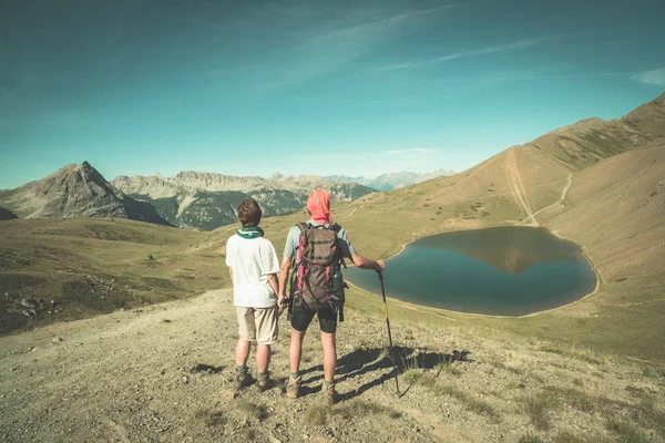 Um par de caminhantes no topo da montanha olhando para o lago azul e picos de montanha. Aventuras de verão nos Alpes. Vista de ângulo largo de cima, imagem tonificada, estilo vintage . — Fotografia de Stock