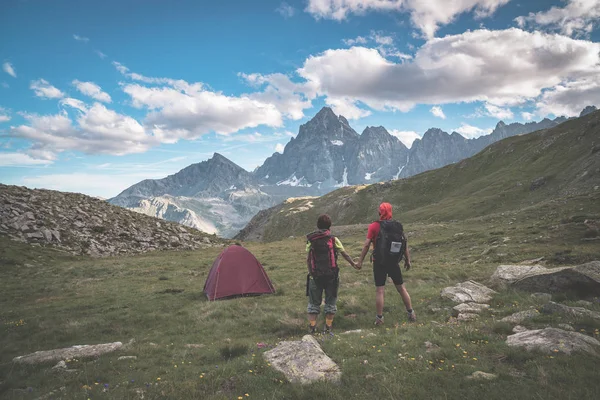 Couple looking at the majestic view of glowing mountain peaks at sunset high up on the Alps. Rear view with camping  tent, focus in the background, toned and filtered image. — Stock Photo, Image