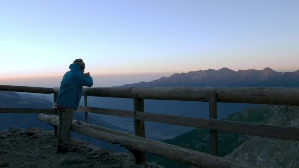 Mann blickt vom Holzbalkon auf die Berge bei Sonnenaufgang und trinkt Heißgetränk aus Becher, hoch oben auf den Alpen. — Stockvideo