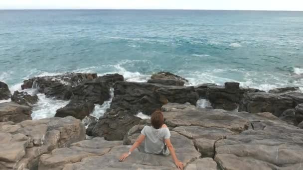 Vista trasera de la mujer sentada en la costa rocosa y mirando el océano agitado desenfocado en el fondo. Cámara lenta, colores naturales . — Vídeos de Stock