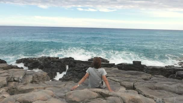 Vista trasera de la mujer sentada en la costa rocosa y mirando el océano agitado desenfocado en el fondo. Cámara lenta, colores naturales . — Vídeo de stock