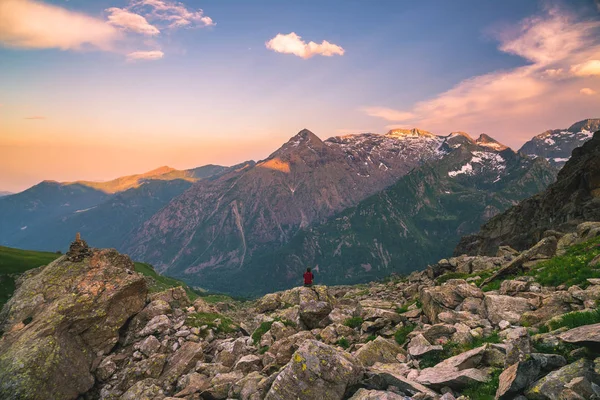 Uma pessoa sentada em terreno rochoso e observando um nascer do sol colorido no alto dos Alpes. Vista de ângulo largo de cima com picos de montanha brilhantes no fundo. Imagem tonificada . — Fotografia de Stock